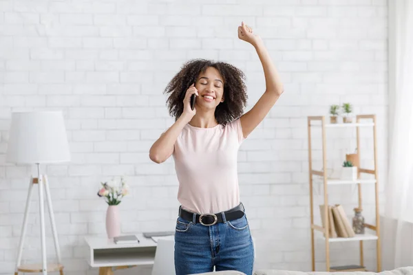 Expresión de felicidad y buenas noticias. Sonriente mujer afroamericana hablando por teléfono y levantando la mano en el interior de la sala de estar durante el autoaislamiento del coronavirus — Foto de Stock