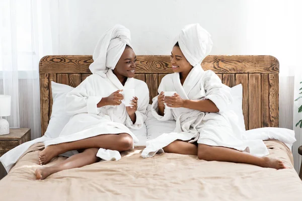 बाथरोब मध्ये आफ्रिकन अमेरिकन स्त्रिया बेडवर बसून, कॉफी पिण्याचे — स्टॉक फोटो, इमेज