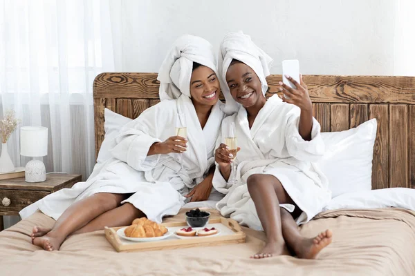 Happy żeńskich przyjaciół w szlafroki siedzi na łóżku, biorąc selfie — Zdjęcie stockowe