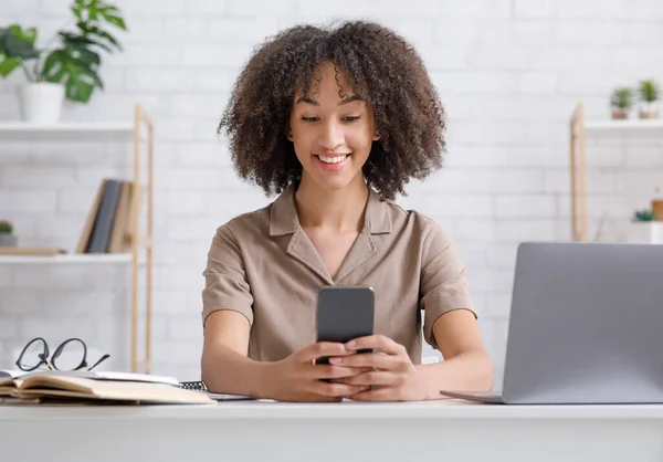 Tecnología moderna para estudiar y trabajar en casa durante la epidemia. Mujer afroamericana sonriente escribiendo en un teléfono inteligente, sentada a la mesa con cuaderno y bloc de notas — Foto de Stock
