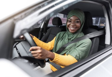 Araba kiralama. Emniyet kemerini takıp araba kullanan neşeli siyah İslamcı kadın.