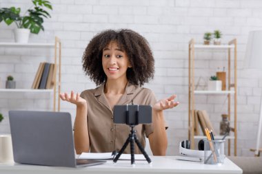 Covid-19 salgını sırasında modern blog yazarlığı ve hobisi. Neşeli Afro-Amerikan kadın takipçiler için video kaydetti ve masadaki bilgisayarlı akıllı telefon web kamerasına baktı.