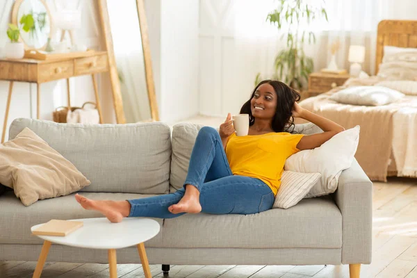 Bella donna nera millenaria che si rilassa sul divano con tazza di bevanda calda a casa — Foto Stock