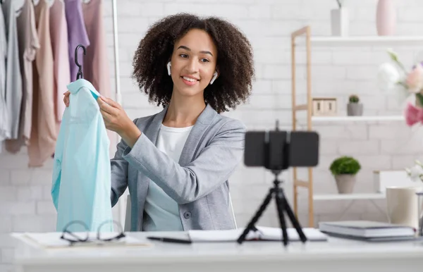 Moda contemporánea y revisión del blog en línea. Mujer afroamericana feliz mostrando ropa en la cámara web del teléfono inteligente en la sala de estar — Foto de Stock