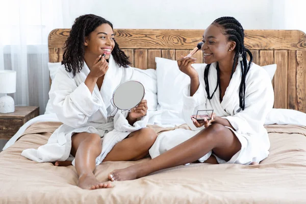 Mulheres negras felizes em roupões de banho colocando maquiagem — Fotografia de Stock