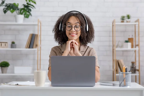 Assista a vídeos, aulas ou webinários remotamente em casa. Mulher americana africana feliz em óculos e fones de ouvido olha para laptop, sentado à mesa no interior da sala de estar moderna — Fotografia de Stock