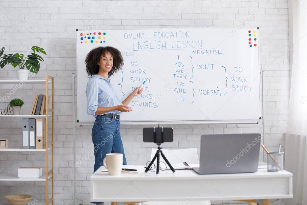 Smiling african american teacher points to white board with rules and records video for students in living room interior with laptop and smartphone