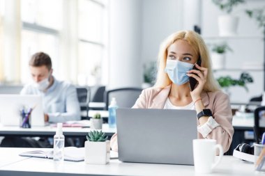Covid-19 karantinasından sonra işine dön. Bin yıllık koruyucu maskeli bir kadın iş yerindeki bilgisayarında modern ofis içi antiseptiklerle telefonla konuşuyor ve daktilo kullanıyor.