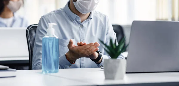 Hand disinfection and health protection from outbreak of covid-19. Guy in protective mask with big bottle of blue antiseptic at table with laptop — Stock Photo, Image