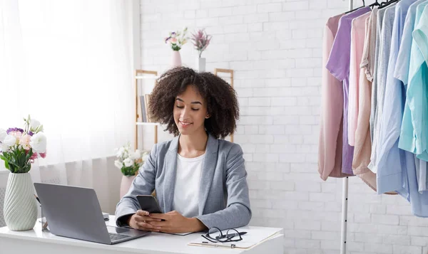 Ótimas avaliações de clientes para pequenas empresas loja de moda on-line. Sorrindo mulher afro-americana lendo no smartphone, sentado à mesa com laptop — Fotografia de Stock