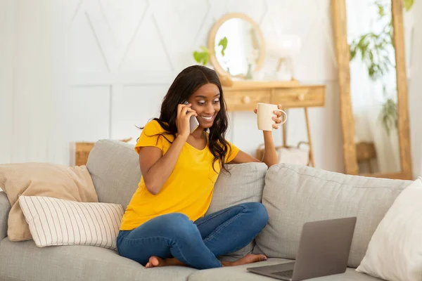 Preciosa mujer negra con bebida caliente usando computadora portátil para el trabajo en línea o estudios mientras habla por teléfono celular en casa —  Fotos de Stock