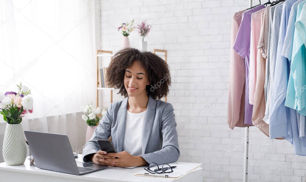 Great reviews from customers to small business online fashion store. Smiling african american woman reading in smartphone, sitting at table with laptop