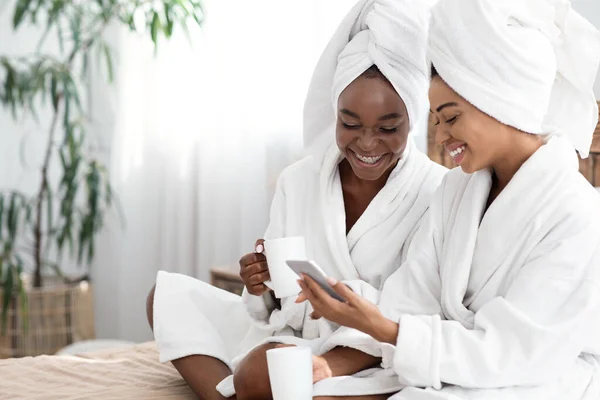 Damas negras felices en albornoces bebiendo café, usando teléfono inteligente — Foto de Stock