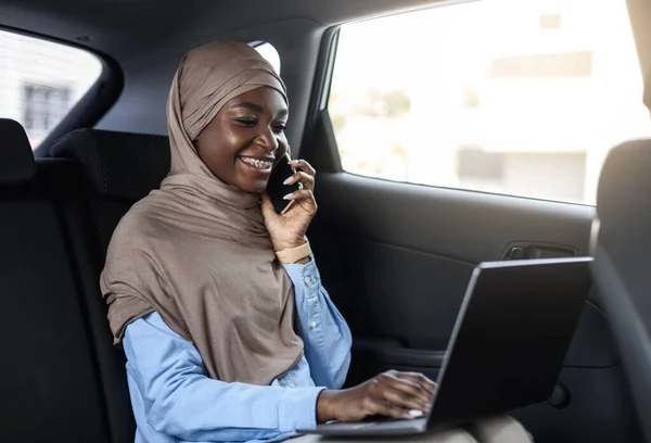 Sonriente mujer de negocios islámica negra hablando por teléfono y utilizando el ordenador portátil en el coche — Foto de Stock