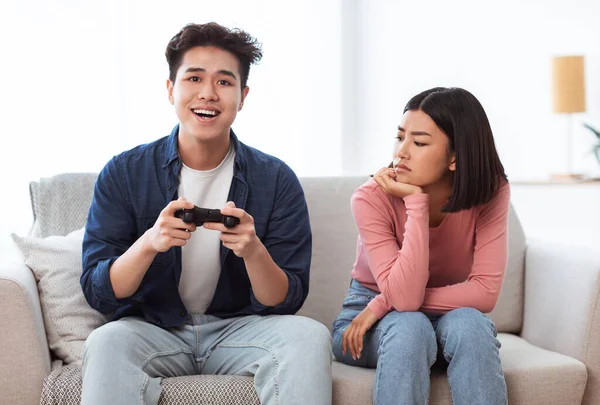Незадоволена азіатська дружина дивиться на чоловіка, граючи в відеоігри в приміщенні — стокове фото