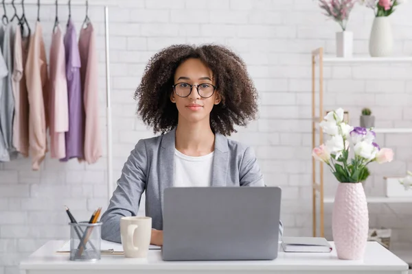 Pequeñas empresas en casa remotamente durante la pandemia de covid-19. Mujer de negocios afroamericana seria diseñadora de moda en gafas sentada en la mesa con computadora portátil en la sala de estar — Foto de Stock