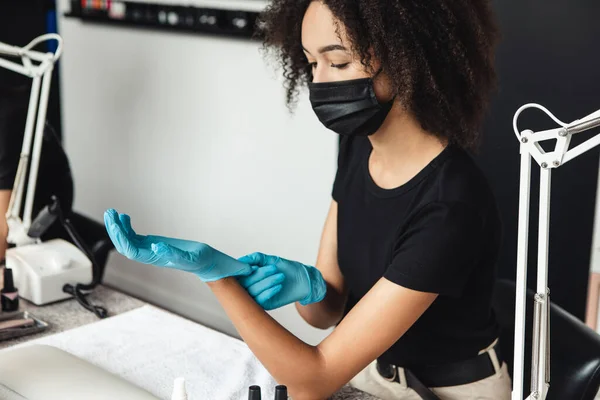 Trabalho de mestre moderno no salão de beleza durante a pandemia de coronavírus. Millennial Africano americano mulher na máscara colocando luvas de borracha no local de trabalho — Fotografia de Stock
