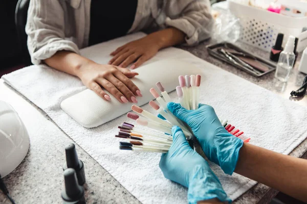 Elija el color de moda en el estudio de belleza. Mujer afroamericana en guantes protectores mostrando muestras de esmalte de uñas al cliente en la mesa con equipo de uñas durante el covid-19 —  Fotos de Stock