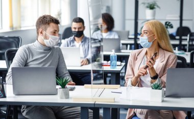 Koruyucu maskeli kadın ve erkek iş yerinde sosyal mesafeyi koruyorlar. Çalışanlar, iç kısımda antiseptik, akıllı telefon ve koruyucu camla masa başında oturmayı tartışıyor