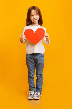 Sarı arka planda büyük kırmızı kalbi tutan anaokulu kızı.