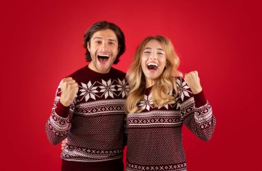 Heyecanlı genç bir çift Noel süveteri içinde çığlık atıyor ve kırmızı arka planda EVET hareketi yapıyor.