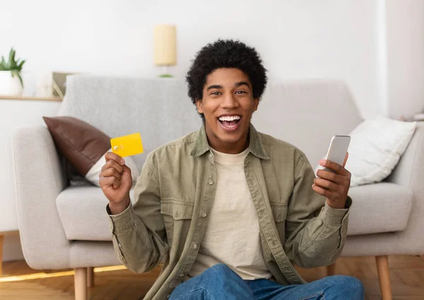 Cybershopping. Eccitato ragazzo adolescente afroamericano con cellulare e carta di credito che ordina merci online da casa — Foto Stock