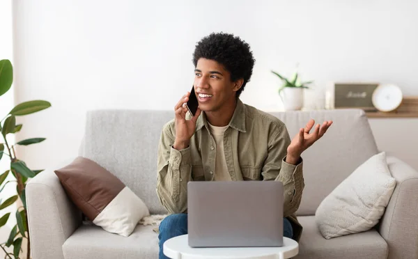 Distance education concept. Happy black student talking on smartphone in front of laptop computer at home — Stock Photo, Image