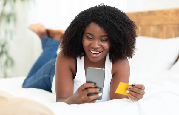 Black lady using phone and credit card in bed