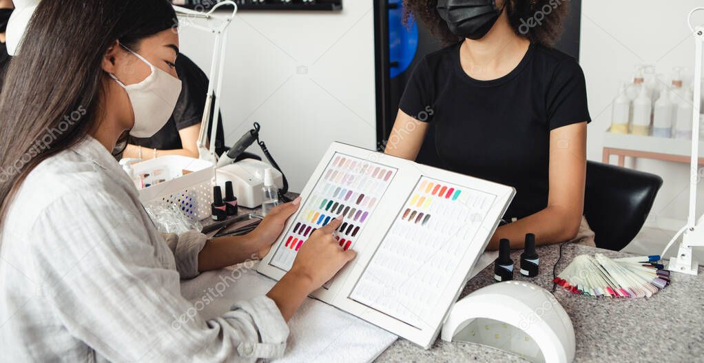 New manicure after lockdown and choice of modern color after quarantine. Asian woman chooses nail polish in catalog with african american master