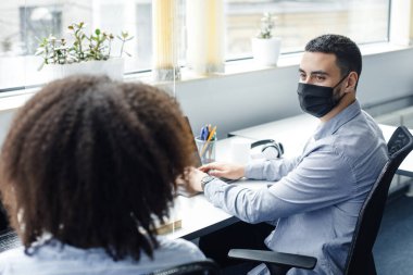 Maskeli adam ve iş yerindeki Afro-Amerikalı kadın ofis içi bilgisayarlarıyla koruyucu camdan konuşuyorlardı.