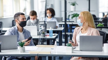 Takım çalışması ve modern teknoloji. Koruyucu maskeli genç adam ve kadın ellerinde akıllı telefonlar, iş yerinde laptoplar ve antiseptiklerle koruyucu cam aracılığıyla konuşuyorlar.