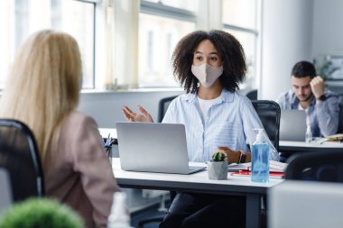 Tecritten sonra müşteri ve çalışanları bulmak. Masasında antiseptik ve dizüstü bilgisayarları olan koruyucu maskeli Afrikalı Amerikalı bir kadın modern ofis içindeki bir bayanla konuşuyor.