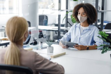 Karantinadan sonra işçi ve boş yer arayın. Afro-Amerikan koruyucu maskeli kadın koruyucu camla sarışın bayana sorular soruyor.