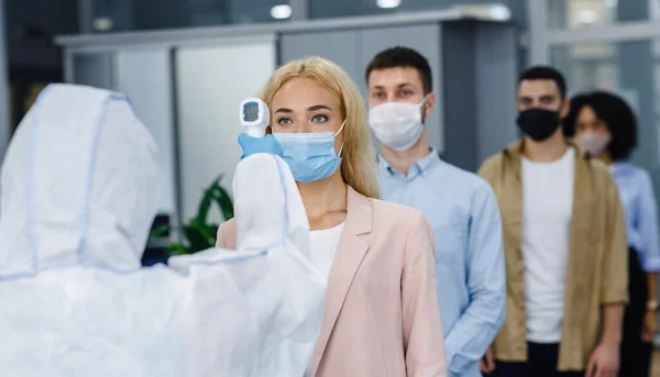 Health control at work and new normal for company. Man in protective suit checks temperature with infrared thermometer at multiracial workers in protective masks