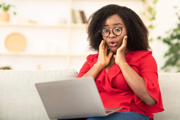 Surprised black woman sitting on couch, using laptop