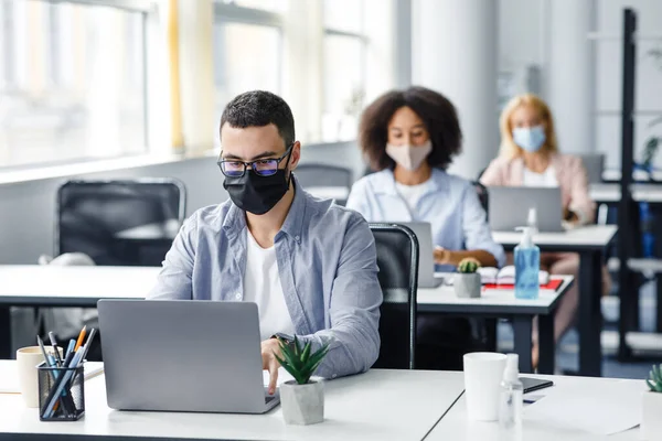 Travail d'équipe en société et retour au travail après la quarantaine covid-19. Focalisé homme millénaire dans des lunettes et masque de protection fonctionne à l'ordinateur portable sur le lieu de travail avec antiseptique — Photo