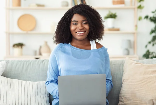 Sort dame sidder på sofaen og arbejder på pc - Stock-foto