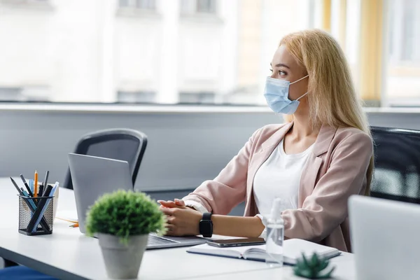 Бизнес должен быть непрерывным, концепция здравоохранения. Блондинка в защитной маске на рабочем месте с ноутбуком и антисептиком и смартфоном смотрит в окно в интерьере — стоковое фото