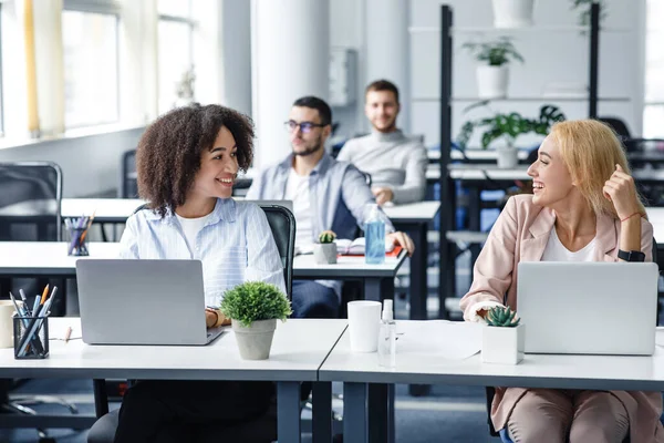 Los trabajadores de las empresas se ocupan de la salud y el distanciamiento social. Sonriente mujer europea y afroamericana en el lugar de trabajo con computadoras portátiles, plantas y antisépticos en la oficina — Foto de Stock