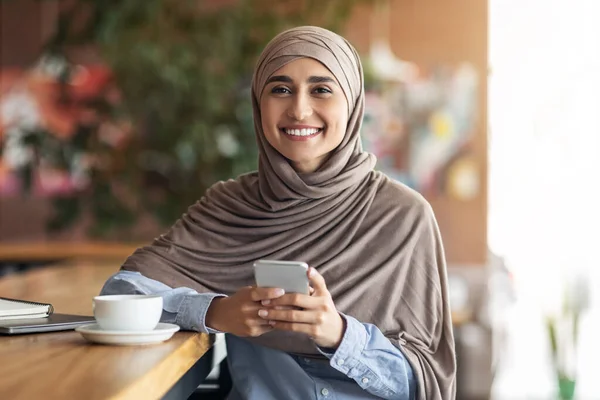 Wanita tersenyum dalam jilbab menggunakan smartphone saat beristirahat di kafe — Stok Foto