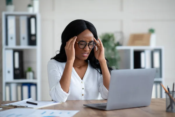 Уставшая чернокожая деловая женщина тысячелетия, страдающая от ужасной головной боли за столом в офисе — стоковое фото