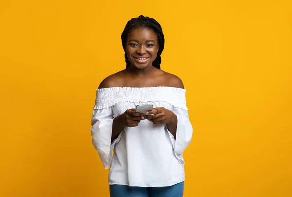 Mobiele communicatie. Portret van gelukkige jonge zwarte vrouw met smartphone, gele achtergrond — Stockfoto