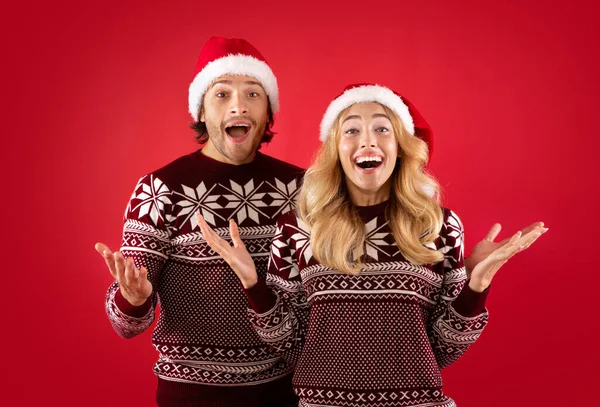 Excited millennial couple in Santa hats and Christmas sweaters over red background — Stock Photo, Image