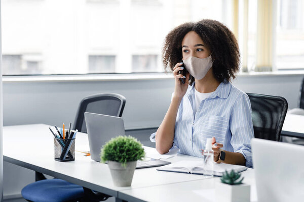 Менеджеры работают с клиентами после карантина. Африканская американка в маске говорит по телефону, сидит на рабочем месте с ноутбуком, антисептик в современном офисе