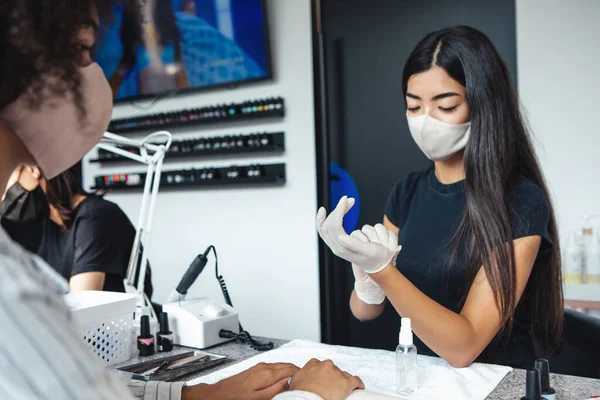 Health care, manicure and social distance. Asian young woman in protective mask wearing rubber gloves for working with nails of african american lady, in beauty salon