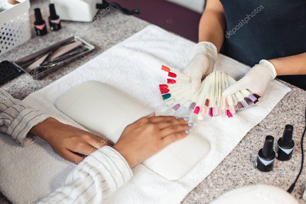 Nail polish color palette. Master in rubber gloves offers samples to african american woman client on table with manicure equipment