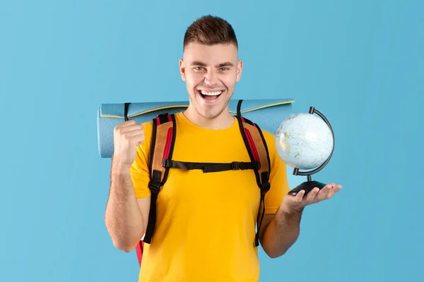 Емоційний чоловік мандрівник з кемпінгом, що тримає глобус і показує жест YES на синьому студійному фоні — стокове фото