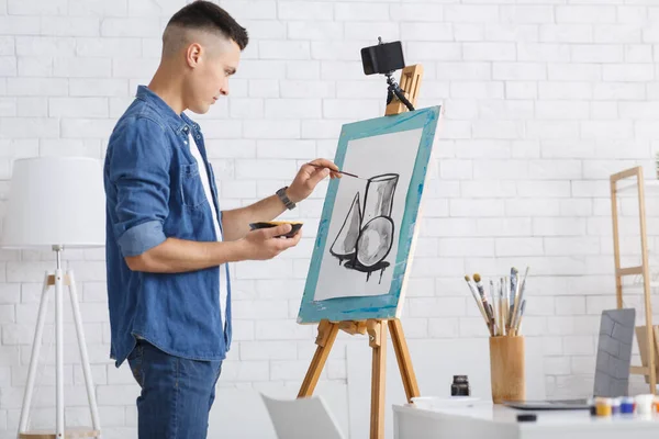 Learn to draw picture and online lesson at home during quarantine. Young guy in denim shirt draws abstract shapes on canvas and watches video on smartphone