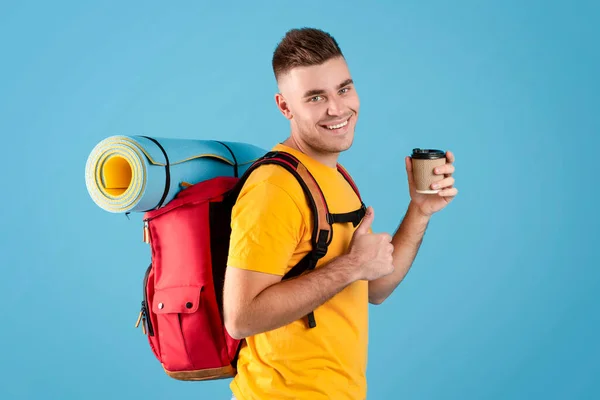 Hombre guapo con equipo turístico sosteniendo el café para llevar y mostrando el gesto hacia arriba pulgar sobre fondo de estudio azul — Foto de Stock