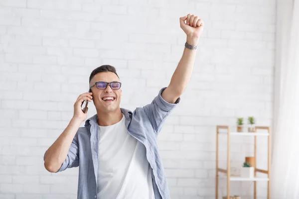 Grand et éloigné travail et gagner à la maison pendant la pandémie. homme heureux dans les lunettes parle au téléphone et exprime des émotions de victoire dans le salon intérieur — Photo